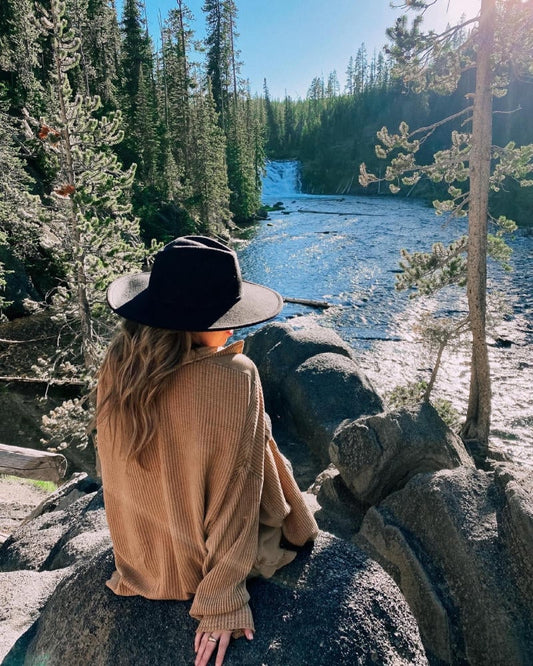 A woman in a felt hat standing by a rocky river, looking out over a forest view in sunlight.